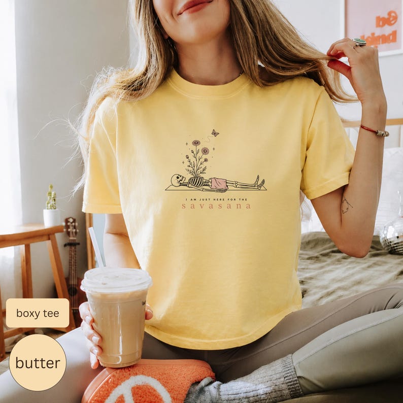 Puede incluir: Una camiseta de manga corta de color amarillo p&aacute;lido con un gr&aacute;fico de l&iacute;neas negras de un esqueleto en pose de yoga, con flores creciendo de su pecho y una peque&ntilde;a mariposa encima. El texto bajo el gr&aacute;fico dice I AM JUST HERE FOR THE SAVASANA.
