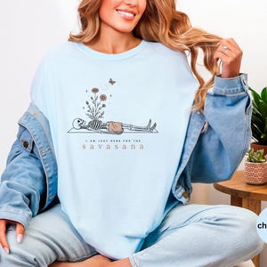 Puede incluir: Una camiseta de cuello redondo azul claro con un gr&aacute;fico de un esqueleto en pose de yoga con flores creciendo de su pecho y una peque&ntilde;a mariposa encima. El texto debajo del esqueleto dice, I AM JUST HERE FOR THE S A V A S A N A.