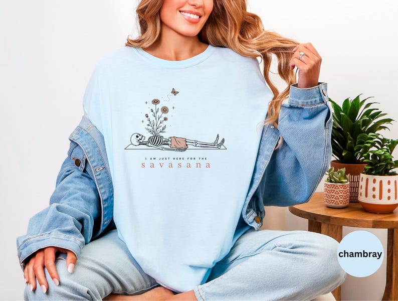 Puede incluir: Una camiseta de cuello redondo azul claro con un gr&aacute;fico de un esqueleto en pose de yoga con flores creciendo de su pecho y una peque&ntilde;a mariposa encima. El texto debajo del esqueleto dice, I AM JUST HERE FOR THE S A V A S A N A.