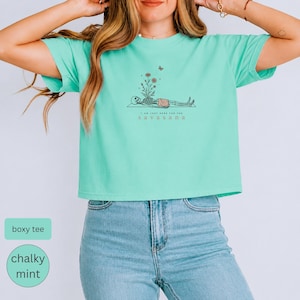 Puede incluir: Una camiseta boxy de color menta tiza con un gr&aacute;fico de un esqueleto en postura de savasana, flores creciendo de su pecho y una peque&ntilde;a mariposa encima. El texto dice I AM JUST HERE FOR THE SAVASANA en una fuente sencilla y clara.