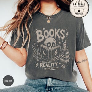 May include: A charcoal gray short-sleeve t-shirt featuring a graphic print of a skull reading a book, a skeleton hand making a peace sign, a candle, and the text "BOOKS because REALITY NEEDS EDITING" in a distressed, off-white ink color.