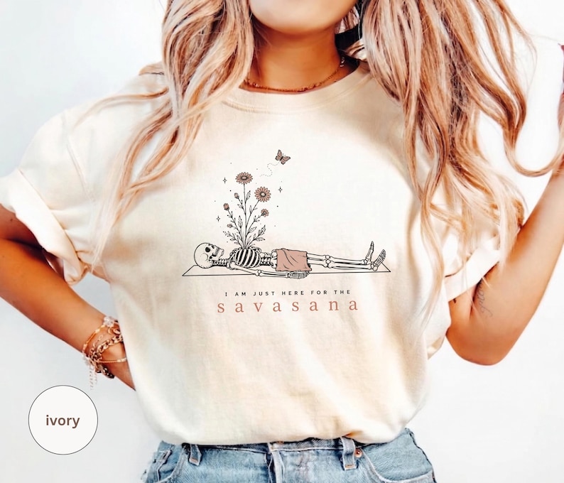 Puede incluir: Una camiseta color marfil con un gr&aacute;fico de l&iacute;neas negras de un esqueleto acostado sobre una esterilla en una postura de yoga. Flores brotan de la caja tor&aacute;cica, una mariposa vuela encima y el texto dice: I AM JUST HERE FOR THE SAVASANA en negro y marr&oacute;n.