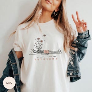 Puede incluir: Una camiseta de color marfil con un gr&aacute;fico de un esqueleto tumbado sobre una esterilla, con flores que brotan de su pecho y una peque&ntilde;a mariposa encima. El texto dice "I AM JUST HERE FOR THE SAVASANA" en letras negras y rojas.