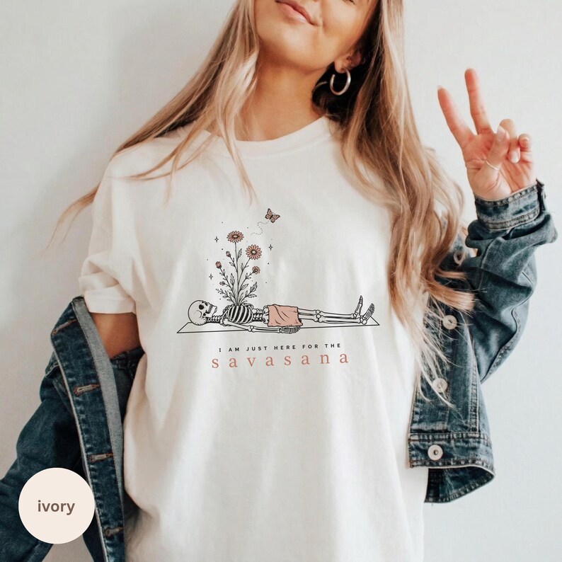 Puede incluir: Una camiseta de color marfil con un gr&aacute;fico de un esqueleto tumbado sobre una esterilla, con flores que brotan de su pecho y una peque&ntilde;a mariposa encima. El texto dice "I AM JUST HERE FOR THE SAVASANA" en letras negras y rojas.