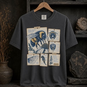 Può includere: Una t-shirt a maniche corte grigio antracite con un'illustrazione scientifica in stile vintage di un bombo. La grafica include diversi diagrammi etichettati dell'insetto, della sua anatomia e di un fiore, presentati su riquadri simili a carta.