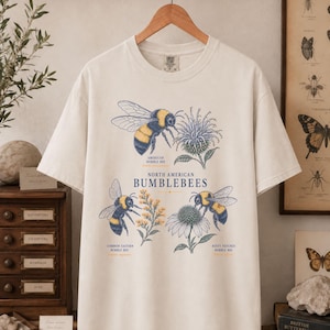May include: An off-white short-sleeve t-shirt featuring a graphic print of three bumblebees and flowers. The text reads North American Bumblebees, American Bumble Bee, Common Eastern Bumble Bee, and Rusty Patched Bumble Bee with scientific names.