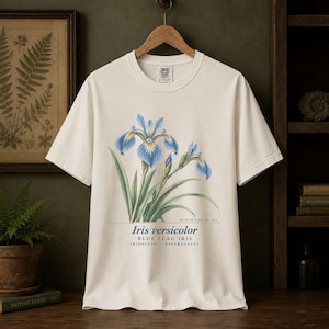 Può includere: Una t-shirt a maniche corte color bianco sporco con un'illustrazione botanica di un fiore di iris blu. Sotto il fiore, il testo recita: Iris versicolor, BLUE FLAG IRIS, IRIDACEAE, ASPARAGALES e REGNUM PLANTAE: 002 in un carattere serif.