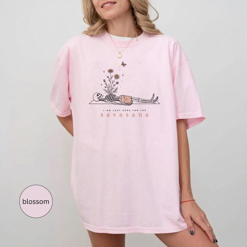 Puede incluir: Una camiseta rosa claro con un gr&aacute;fico de un esqueleto tumbado con flores creciendo en su pecho. Sobre el esqueleto hay una peque&ntilde;a mariposa. El texto "I AM JUST HERE FOR THE SAVASANA" est&aacute; impreso en negro y naranja debajo del gr&aacute;fico.