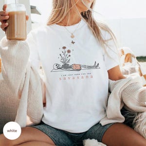 Puede incluir: Una camiseta blanca con un gr&aacute;fico de l&iacute;neas negras de un esqueleto en pose de yoga, con flores creciendo de su pecho y una peque&ntilde;a mariposa encima. El texto "I AM JUST HERE FOR THE SAVASANA" est&aacute; impreso en letras negras y naranjas.