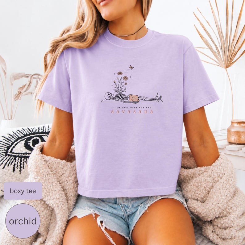 Puede incluir: Una camiseta de corte cuadrado en color violeta claro con un gr&aacute;fico de un esqueleto tumbado en una esterilla de yoga, flores que brotan de su pecho, una peque&ntilde;a mariposa y el texto I AM JUST HERE FOR THE SAVASANA en letras negras y naranjas.