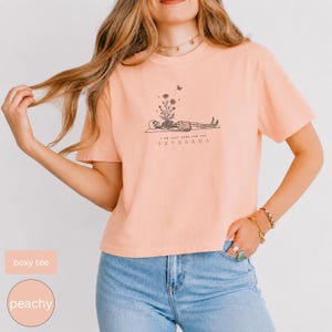 Puede incluir: Una camiseta de corte cuadrado color melocot&oacute;n con un gr&aacute;fico de l&iacute;neas negras de un esqueleto tumbado en una esterilla de yoga, con flores brotando de su pecho y una peque&ntilde;a mariposa. El texto debajo dice I AM JUST HERE FOR THE SAVASANA.