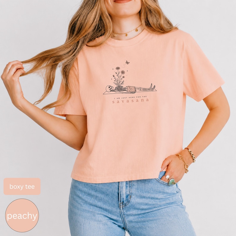 Puede incluir: Una camiseta de corte cuadrado color melocot&oacute;n con un gr&aacute;fico de l&iacute;neas negras de un esqueleto tumbado en una esterilla de yoga, con flores brotando de su pecho y una peque&ntilde;a mariposa. El texto debajo dice I AM JUST HERE FOR THE SAVASANA.