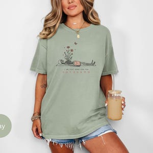 Puede incluir: Una camiseta de manga corta color verde salvia con un gr&aacute;fico de un esqueleto acostado sobre una esterilla con flores creciendo de su pecho. El texto dice I AM JUST HERE FOR THE SAVASANA. El dise&ntilde;o incluye una peque&ntilde;a mariposa y flores.