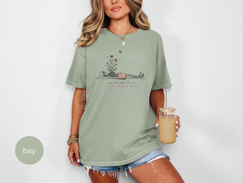 Puede incluir: Una camiseta de manga corta color verde salvia con un gr&aacute;fico de un esqueleto acostado sobre una esterilla con flores creciendo de su pecho. El texto dice I AM JUST HERE FOR THE SAVASANA. El dise&ntilde;o incluye una peque&ntilde;a mariposa y flores.