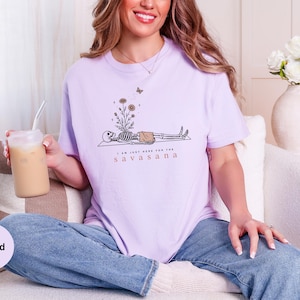 Puede incluir: Una camiseta de manga corta de color morado claro con un gr&aacute;fico de un esqueleto tumbado con flores creciendo en el pecho y una mariposa encima. El texto "I AM JUST HERE FOR THE SAVASANA" est&aacute; impreso en letras negras y marrones.