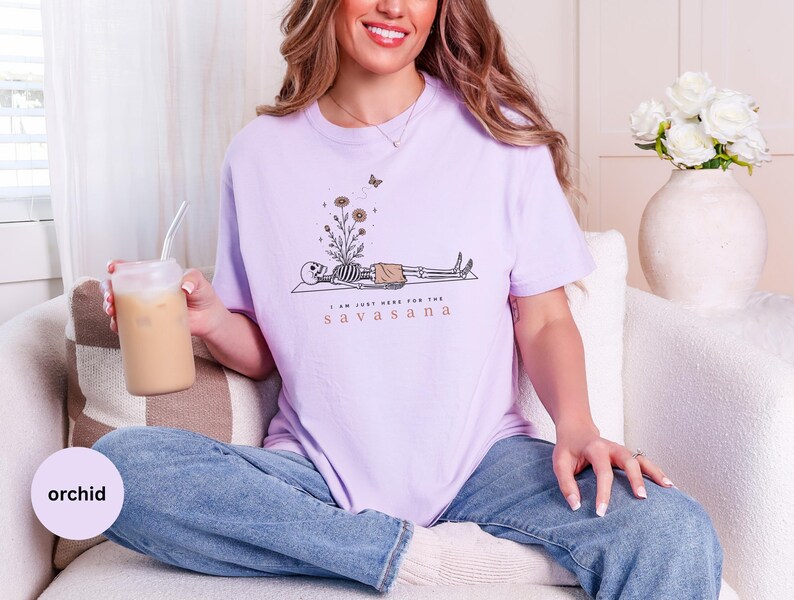 Puede incluir: Una camiseta de manga corta de color morado claro con un gr&aacute;fico de un esqueleto tumbado con flores creciendo en el pecho y una mariposa encima. El texto "I AM JUST HERE FOR THE SAVASANA" est&aacute; impreso en letras negras y marrones.
