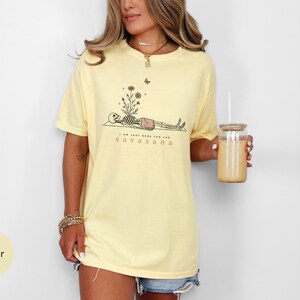 Puede incluir: Una camiseta de manga corta color amarillo mantequilla con un gr&aacute;fico de un esqueleto en postura de yoga, flores creciendo de su pecho y una peque&ntilde;a mariposa encima. El texto dice I AM JUST HERE FOR THE SAVASANA en color marr&oacute;n oscuro.