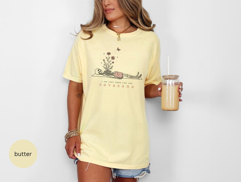 Puede incluir: Una camiseta de manga corta color amarillo mantequilla con un gr&aacute;fico de un esqueleto en postura de yoga, flores creciendo de su pecho y una peque&ntilde;a mariposa encima. El texto dice I AM JUST HERE FOR THE SAVASANA en color marr&oacute;n oscuro.