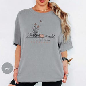Puede incluir: Una camiseta gris de manga corta con un gr&aacute;fico de un esqueleto tumbado con flores creciendo en su pecho y una mariposa encima. El texto dice I AM JUST HERE FOR THE SAVASANA, con la palabra SAVASANA impresa en un color naranja apagado.