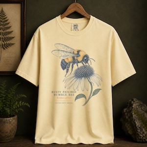 May include: A pale yellow short-sleeve t-shirt featuring a graphic illustration of a bumblebee on a flower. The text reads RUSTY PATCHED BUMBLE BEE, Bombus affinis, and EASTERN NORTH AMERICA. The shirt has a crew neck and a Comfort Colors brand label.