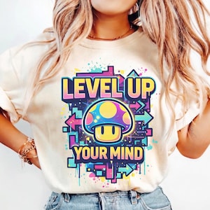 Könnte beinhalten: Cremefarbenes T-Shirt mit einem farbenfrohen Grafikdesign, das einen Pilz und den Text "LEVEL UP YOUR MIND" zeigt. Die Grafik enthält Pfeile und geometrische Formen in Blau-, Rosa-, Gelb- und Lilatönen. Das T-Shirt hat kurze Ärmel.