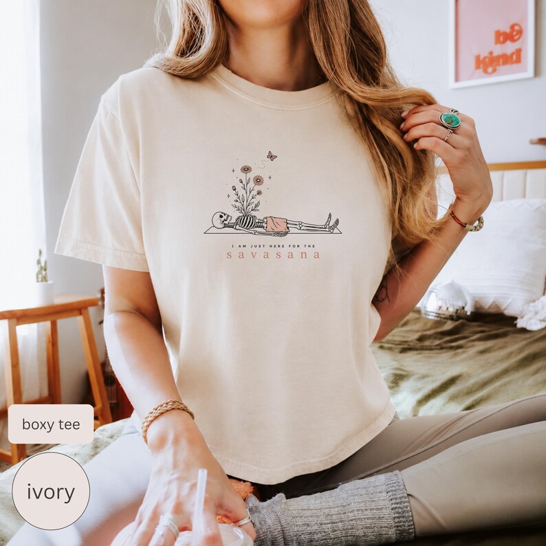 Puede incluir: Una camiseta de manga corta color marfil con un gr&aacute;fico de un esqueleto tumbado en una esterilla de yoga, con flores creciendo de su pecho y una peque&ntilde;a mariposa encima. El texto bajo el gr&aacute;fico dice: "I AM JUST HERE FOR THE SAVASANA."