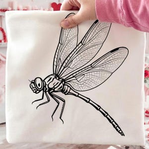 May include: A white tote bag with a black and white dragonfly design. The dragonfly is illustrated with detailed wings and a thin body. The bag is held by a person.
