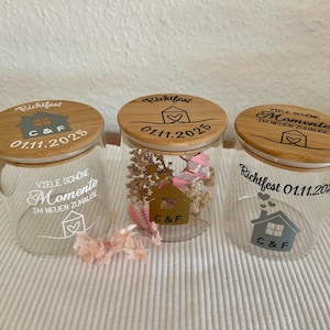 Könnte beinhalten: Drei Glasbehälter mit Bambusdeckeln, jeweils mit individuellem Text und Grafiken. Auf den Gläsern stehen die Wörter "Richtfest" und "Viele Schone Momente", eine Hausgrafik und das Datum 01.11.2025. Ein Glas ist mit Trockenblumen gefüllt.