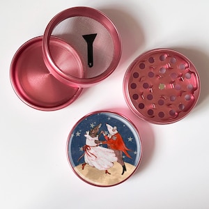 May include: A disassembled pink herb grinder with a screen, a scraper, and a lid. The bottom piece features a magnet. A decorative bottom piece shows a dancing couple with a starry background.