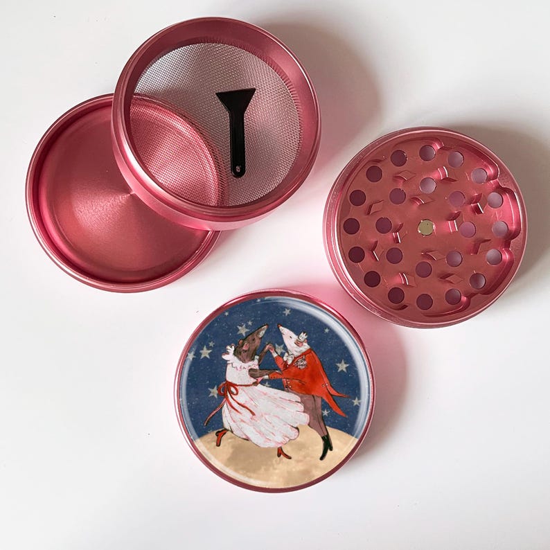 May include: A disassembled pink herb grinder with a screen, a scraper, and a lid. The bottom piece features a magnet. A decorative bottom piece shows a dancing couple with a starry background.