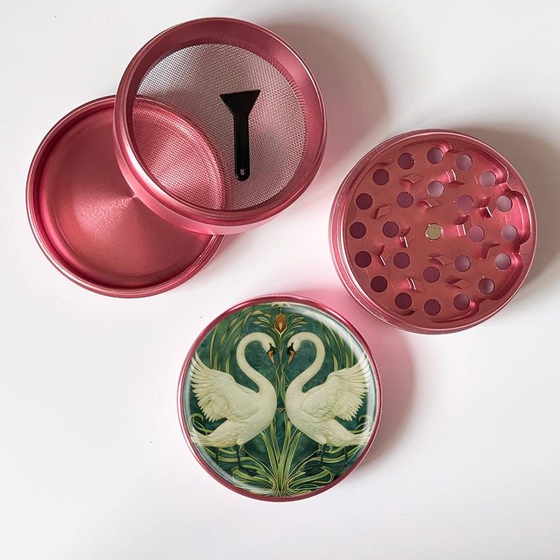 May include: A disassembled pink herb grinder with a screen, a small black scraper, and a decorative lid. The lid features a design of two white swans facing each other against a green and gold background.