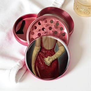 May include: A pink, disassembled herb grinder with a black scraper. The grinder's top component features a circular image of a woman in a red dress and long gloves. The grinder is on a white surface.
