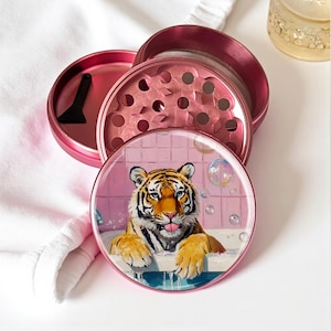 May include: A circular pink metal herb grinder featuring a printed illustration of a tiger in a bathtub on the lid. The grinder is disassembled into three parts, revealing internal grinding teeth and a small black plastic scraping tool.