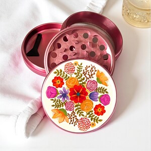 May include: A pink metal herb grinder with a floral design. The grinder is disassembled, revealing the components, including a small black scraper. The floral design features a variety of colorful flowers and leaves on a white background.