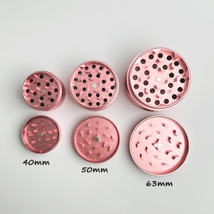 May include: Six rose-gold metal grinders in various sizes. The grinders are round with multiple holes and a magnetic center. The sizes are labeled as 40mm, 50mm, and 63mm. The grinders are designed for herbs.