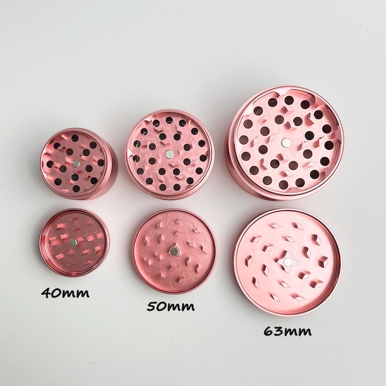May include: Six rose-gold metal grinders in various sizes. The grinders are round with multiple holes and a magnetic center. The sizes are labeled as 40mm, 50mm, and 63mm. The grinders are designed for herbs.