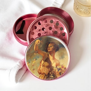 May include: A pink, four-piece herb grinder with a vintage art print on the lid. The art features a woman holding flowers. The grinder includes a small black scraper. The grinder is on a white surface.