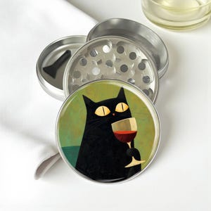 May include: A silver metal herb grinder with a multi-piece design and a small black scraper tool. The top lid features a circular illustration of a black cat with large yellow eyes holding a stemmed glass filled with red wine.