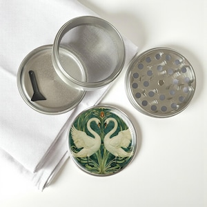 May include: A disassembled herb grinder in silver metal, with a mesh screen, a small black scraper, and a decorative base. The base has a circular design with two white swans facing each other on a green and gold background.