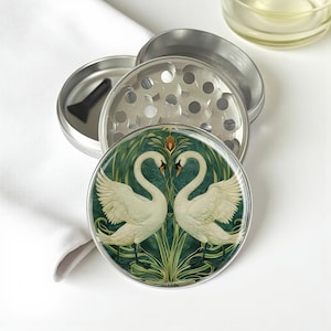 May include: A silver metal herb grinder with a decorative top featuring two white swans facing each other against a green and teal background. The grinder is partially disassembled, showing its components.