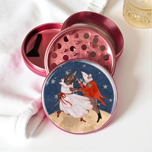 May include: A pink, disassembled grinder with a whimsical illustration of two dancing rats on the top. The grinder includes a small black scraper. The background is a starry night scene.