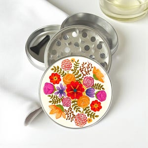 May include: A silver metal herb grinder with a floral design. The grinder is disassembled, revealing the components. The top piece features a colorful floral pattern with red, orange, and purple flowers. A black scraper is included.