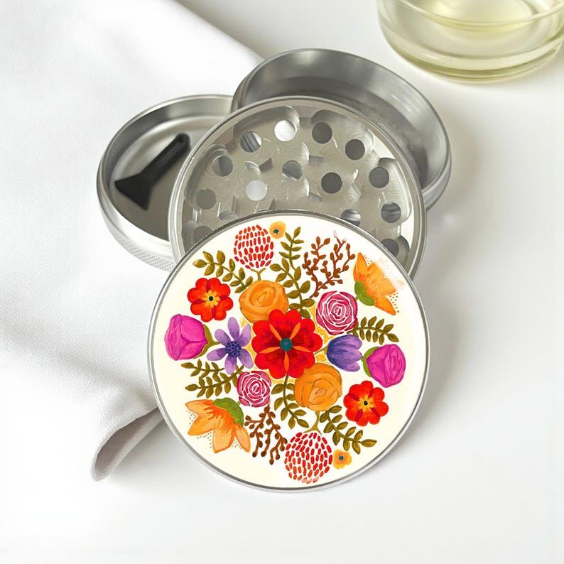 May include: A silver metal herb grinder with a floral design. The grinder is disassembled, revealing the components. The top piece features a colorful floral pattern with red, orange, and purple flowers. A black scraper is included.