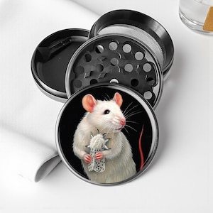 May include: A black and silver herb grinder with a white rat illustration on the lid. The rat is holding a small, patterned object against a black background. The grinder is disassembled, showing its various components.