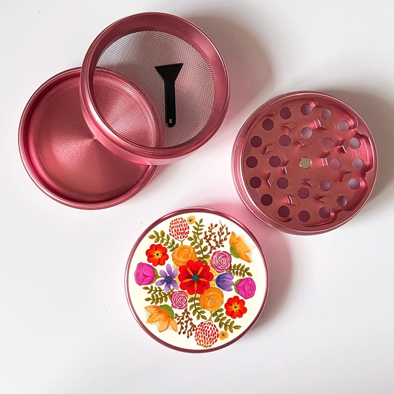 May include: A disassembled pink metal herb grinder with a floral design on the bottom piece. The grinder includes a mesh screen, a small black scraper, and a top piece with grinding teeth.