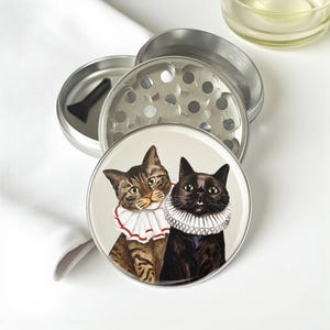 May include: A silver metal herb grinder with a two-cat design. One cat is brown tabby, the other is black, both wearing ruffled collars. The grinder is disassembled, showing the grinding plate and a small black scraper.