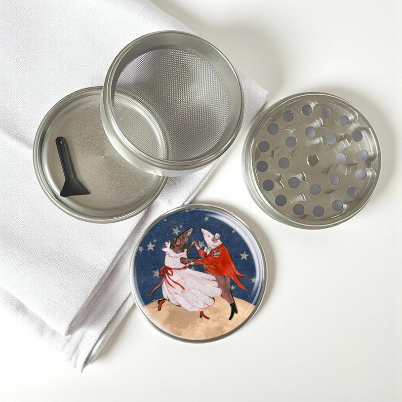 May include: A disassembled silver metal herb grinder with a mesh screen, a small black scraper, and a lid featuring a dancing couple illustration on a starry background. The grinder is on a white cloth.