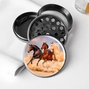 May include: A black metal herb grinder with a three-part design. The top component features a mesh screen and a series of holes. The bottom component has a circular image of three horses running through a sandy landscape.