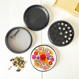 May include: A four-piece black herb grinder with a floral design on the bottom piece. The grinder is disassembled, showing the top, the grinding section, the sifting screen with a small scraper, and the bottom piece. A small pile of herbs is visible.