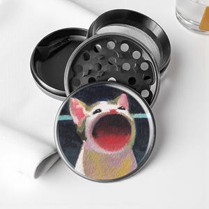 May include: A black and silver herb grinder with a cat illustration on the lid. The cat has a wide open mouth and is painted in shades of white, yellow, and red. The grinder is disassembled, showing the different parts.